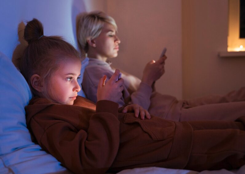 Mother and her young daughter in a bed at night watching their smartphones
