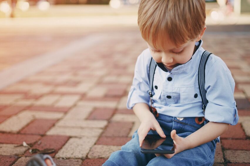 A toddler sitting on the ground and looking at a smartphone instead of playing