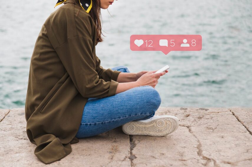 Woman sitting on the ground by the sea checking her social media for likes and messages