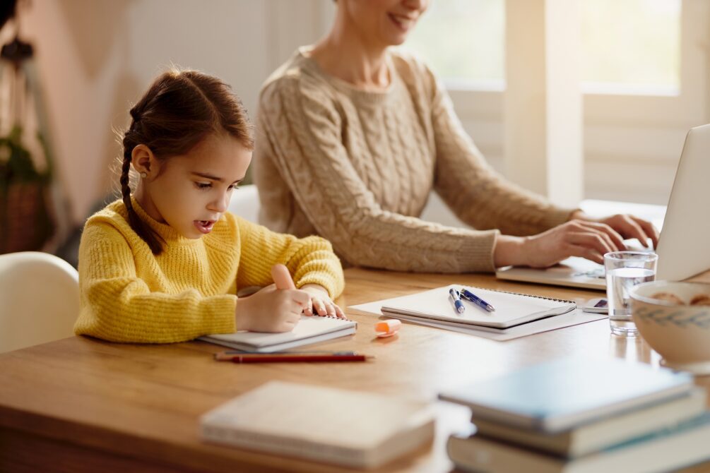 Little girl doing homework while mother is working on a computer at home.
