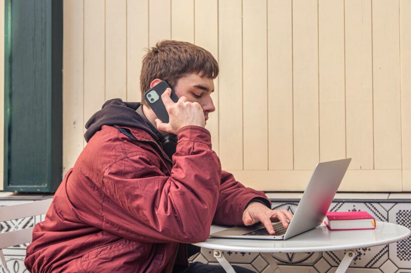 A young man, alone, talking on the phone and using laptop at the same time.