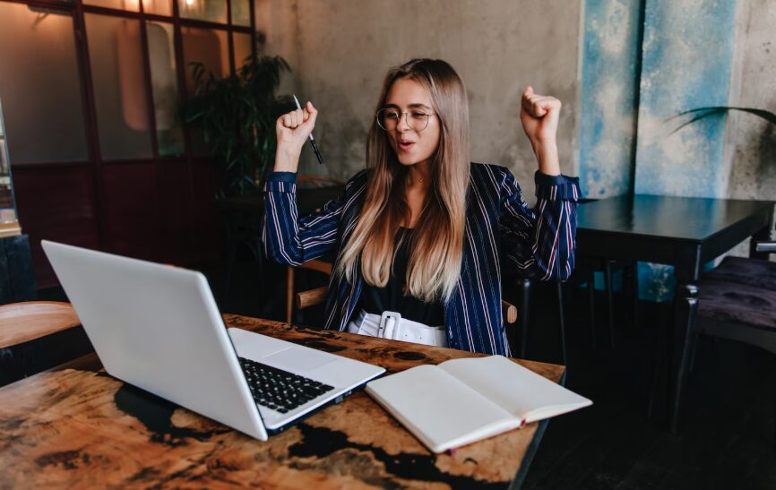 A young woman smiling and cheering while looking at her laptop.
