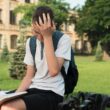 A boy sitting in front of a university holding his head tired of reading a book