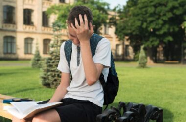 A boy sitting in front of a university holding his head tired of reading a book