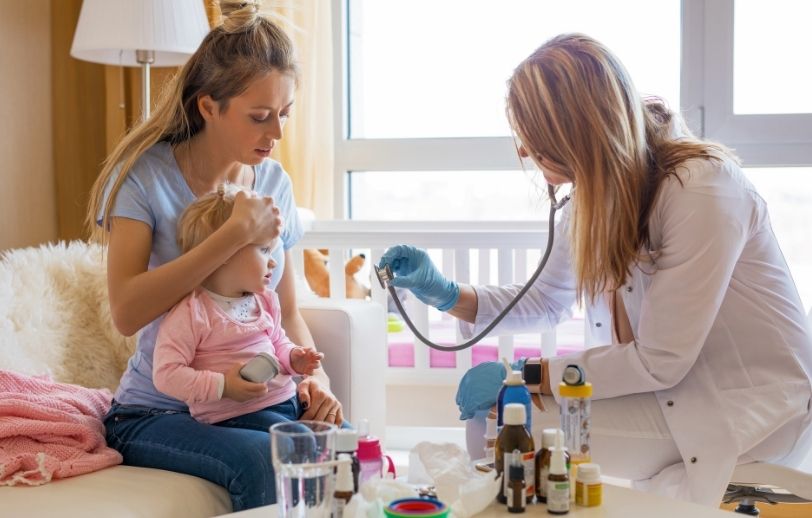 Mother holding a sick baby while being examined by a doctor.
